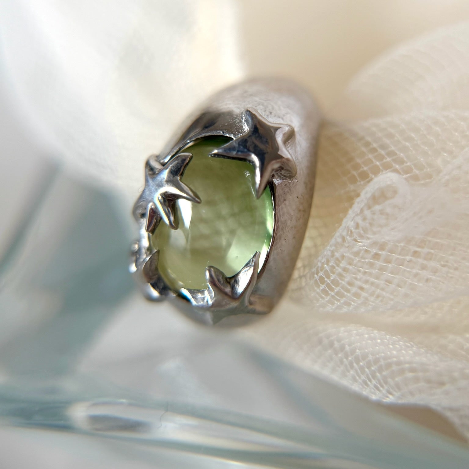 Close-up of a silver ring featuring a light green round stone held by star-shaped prongs on fabric.
