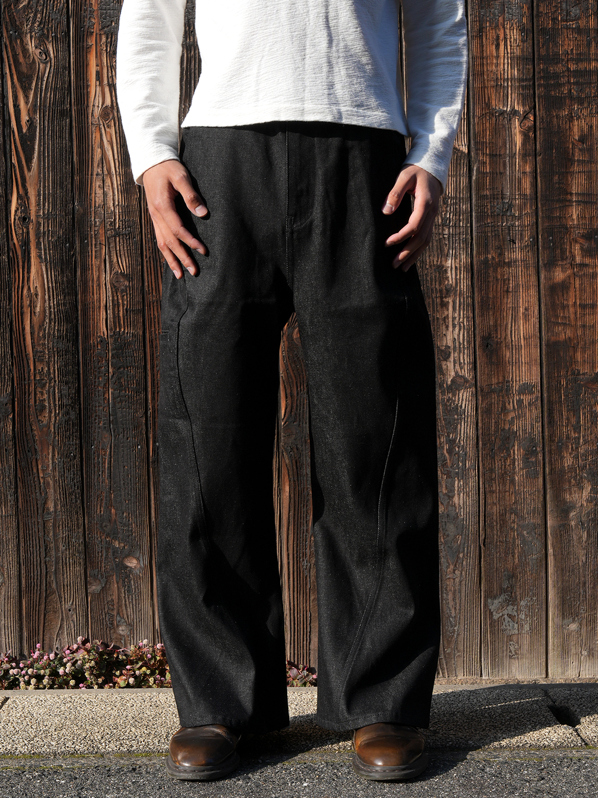 Front view of the black Twisted Raw Jeans worn by a model standing against a wooden wall.