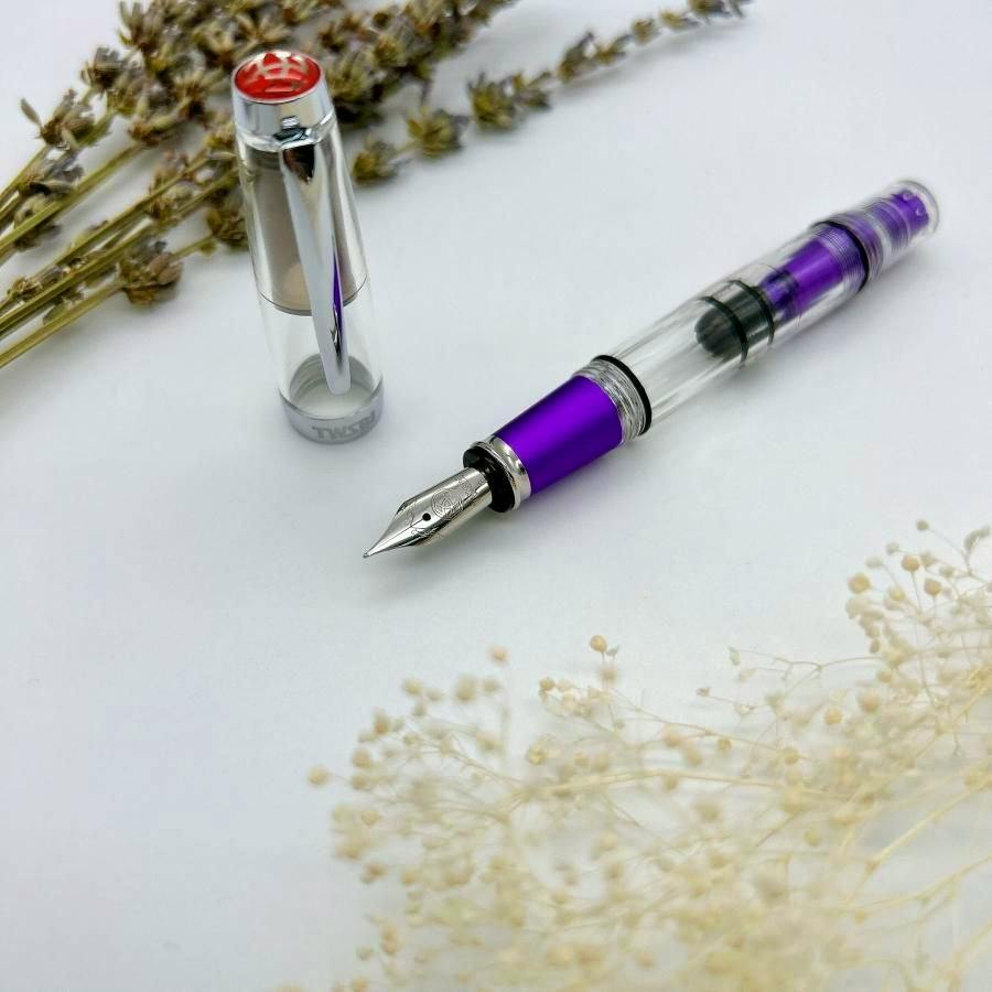 A TWSBI DIAMOND Mini AL Grape fountain pen shown uncapped, with the purple grip section and clear barrel visible and the cap placed beside it on a white background with dried flowers.