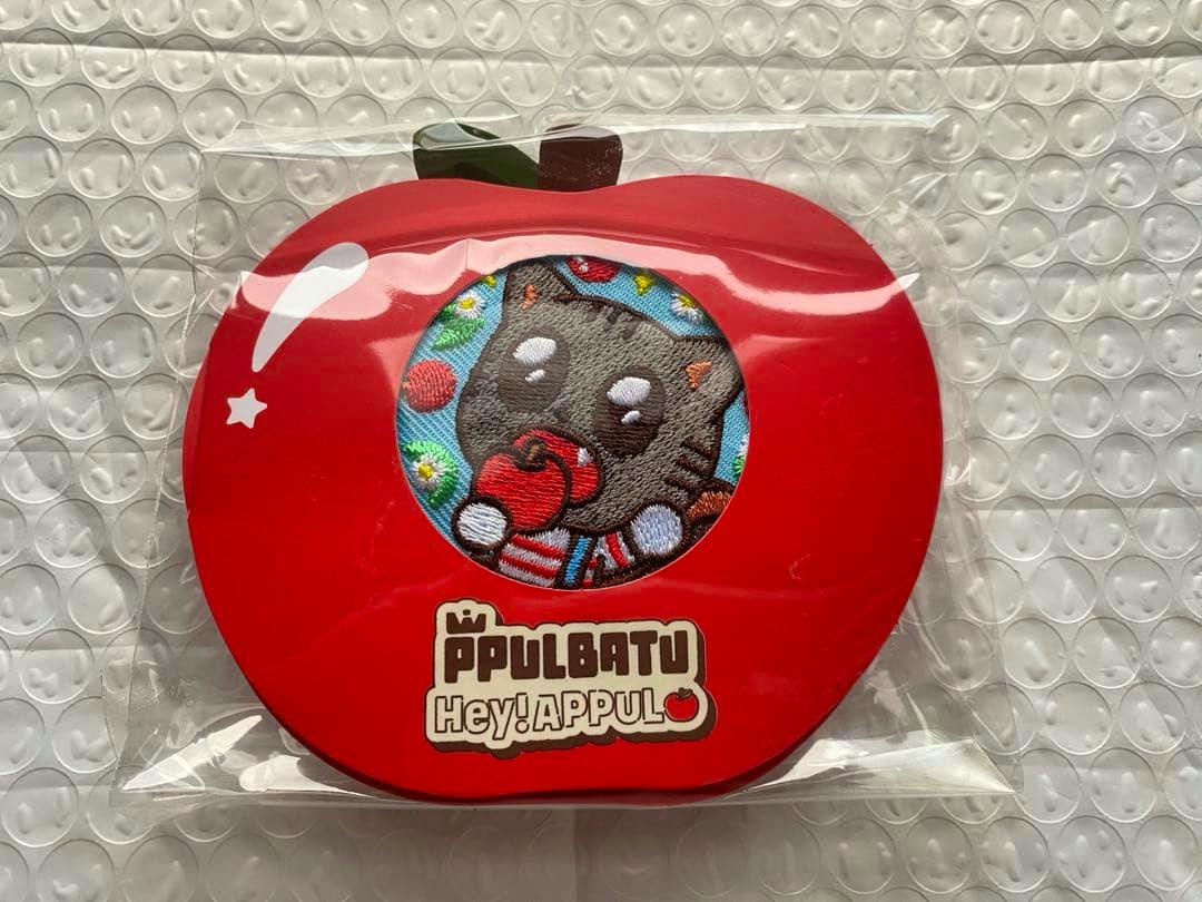 Front of red apple-shaped packaging containing a can badge with an embroidered cat holding an apple visible through a round window.