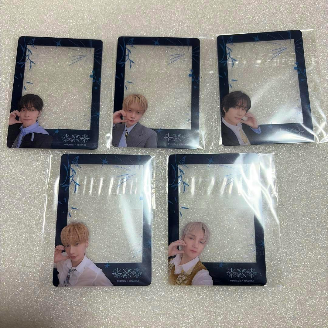 Five clear TXT photocard frames in plastic sleeves are arranged on a sparkly surface, each showing a member portrait in the lower corner.