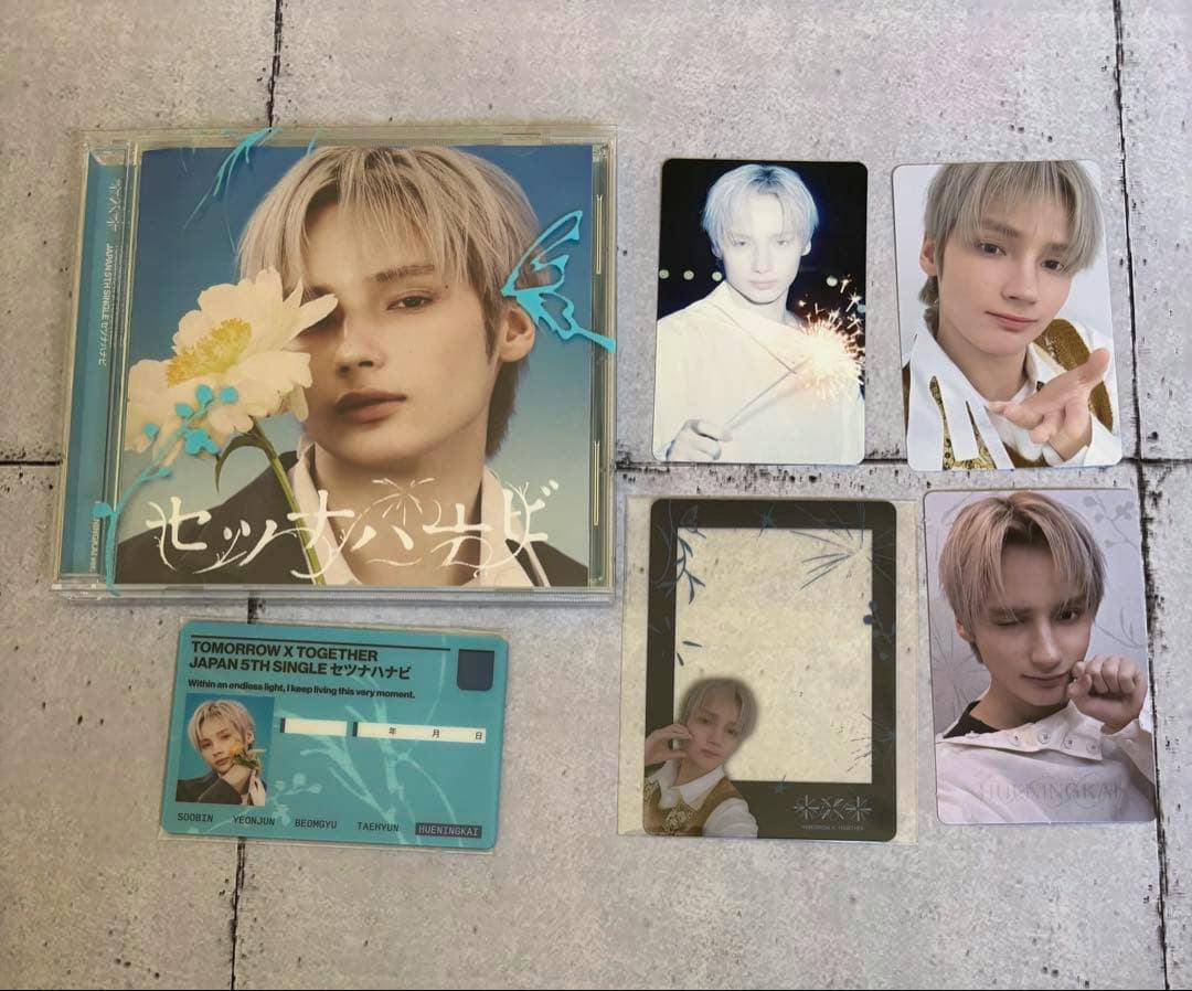 A flat lay of the TXT CD single case with several member photo cards and collectible cards arranged beside it.