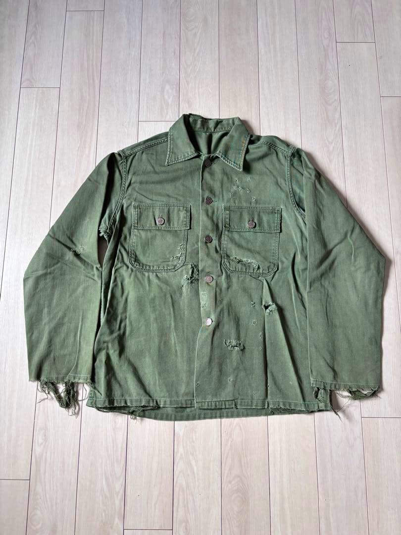 Front view of a men's U.S. Army green cotton utility shirt with button closure, two chest pockets, and visible distressing and frayed hems.