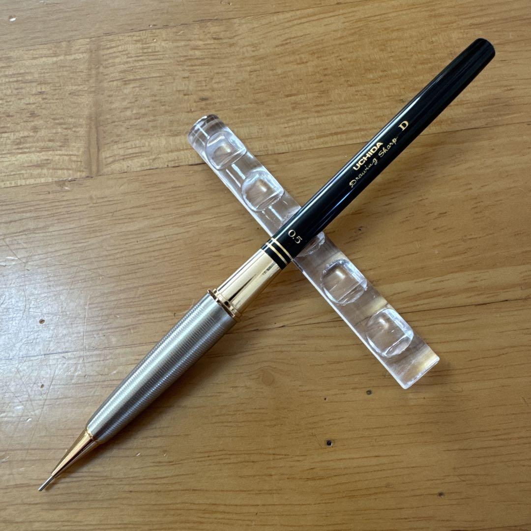UCHIDA D-type 0.5 mechanical pencil with a metal grip shown on a clear stand on a wooden surface.