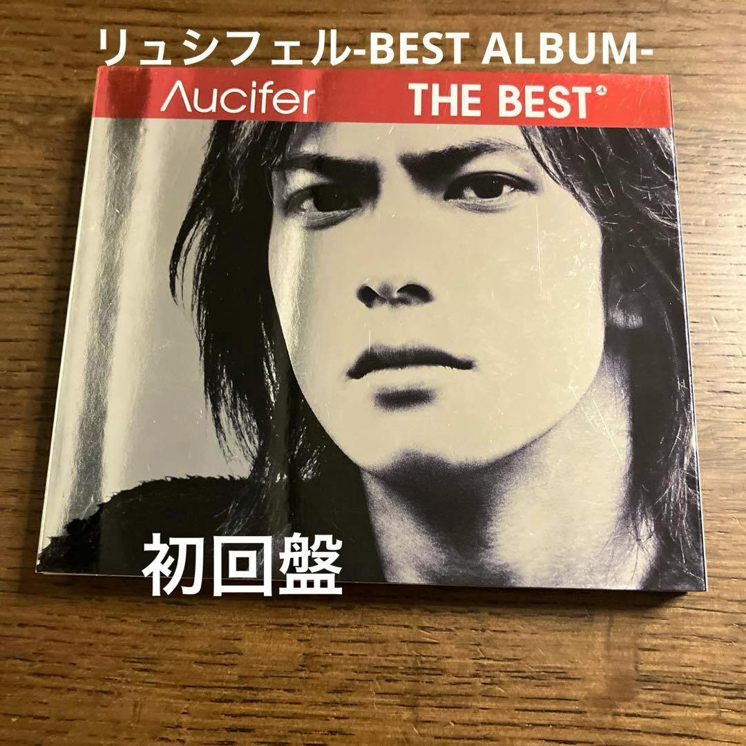 Front cover of the Λucifer リュシフェル 『THE BEST』 初回盤 CD showing a black-and-white close-up portrait with a red header.