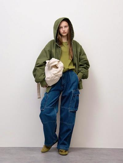 A model wearing blue denim wide cargo pants with large side cargo pockets, paired with a green hooded jacket and holding a beige bag.