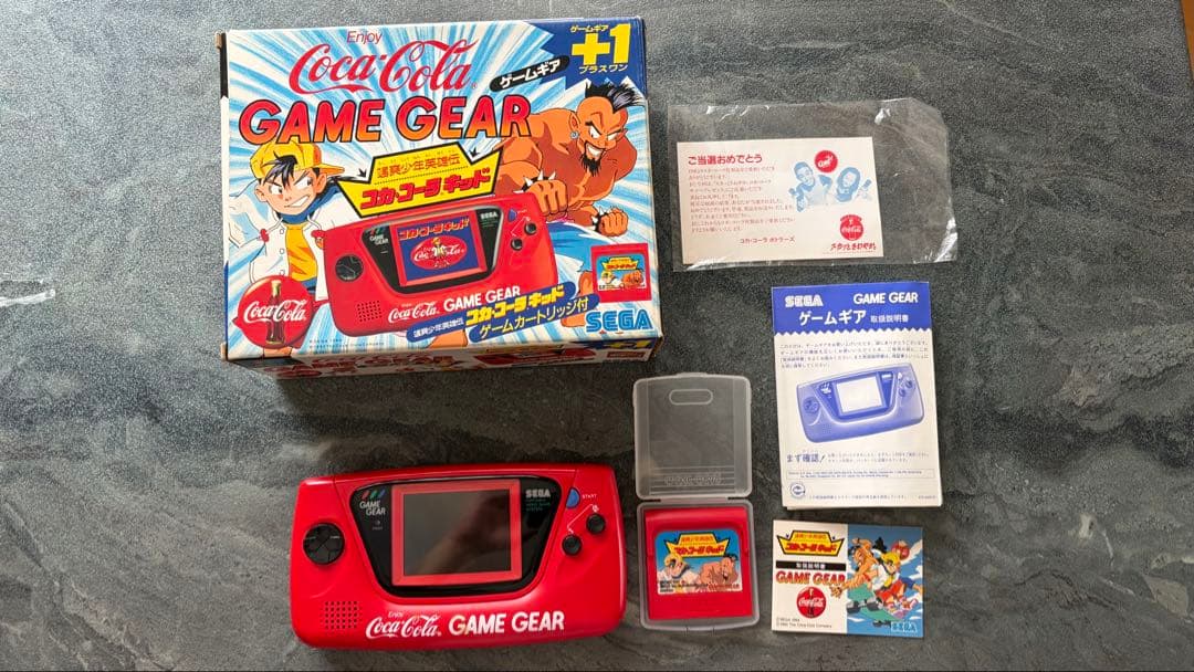 Red Coca‑Cola Sega Game Gear console with its Coca‑Cola box, game cartridge, manuals, and a clear cartridge case laid out on a surface.
