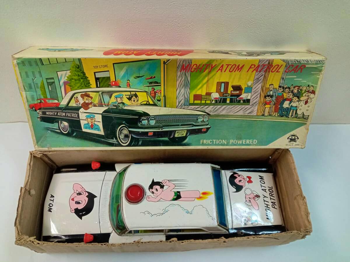 Tin Mighty Atom Patrol Car toy and its illustrated box shown inside an open cardboard box.