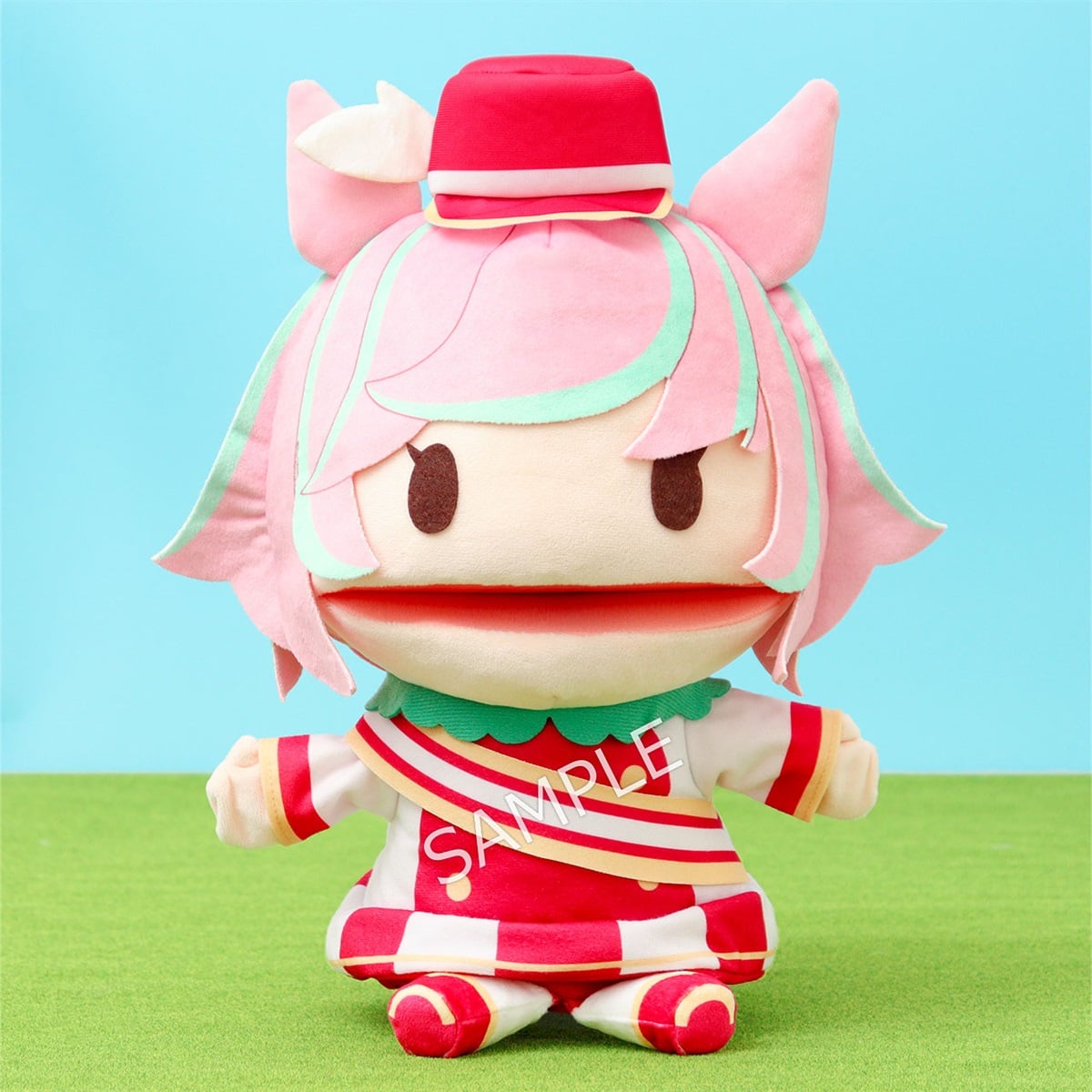Front-facing photograph of the マルシュロレーヌ (Marsh) puppet plush showing pink hair, a small red hat, striped red-and-white outfit, and open mouth, seated on a green surface against a blue background.