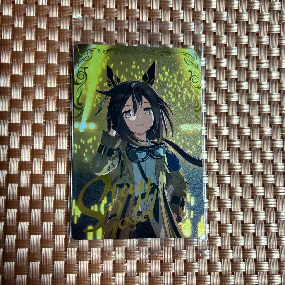 A Stay Gold (ウマ娘) photocard in a clear plastic sleeve placed on a woven placemat.