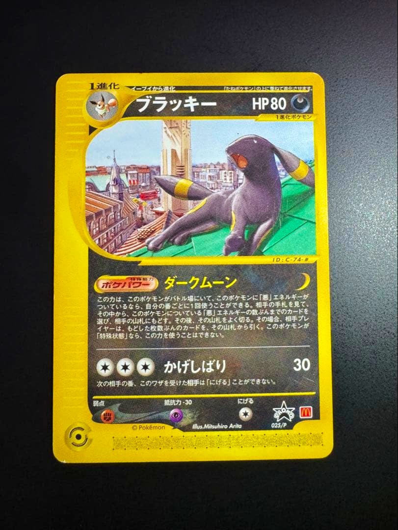 Photo of the Japanese ブラッキー (Umbreon) promo Pokémon trading card 025/P with a yellow border lying on a dark surface.