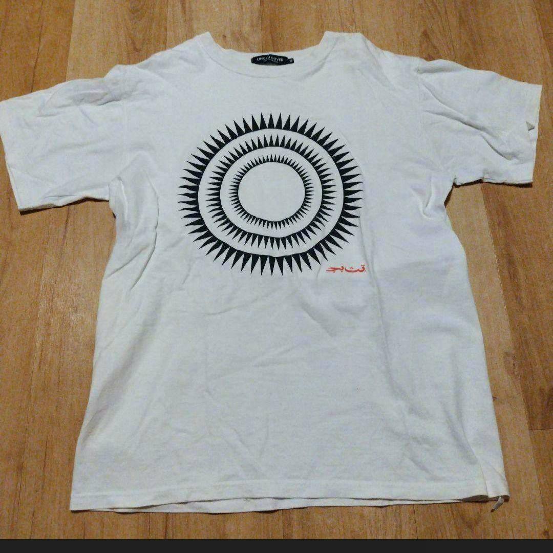 A white T-shirt laid flat on a wooden floor showing a black concentric spiked circle print on the front.