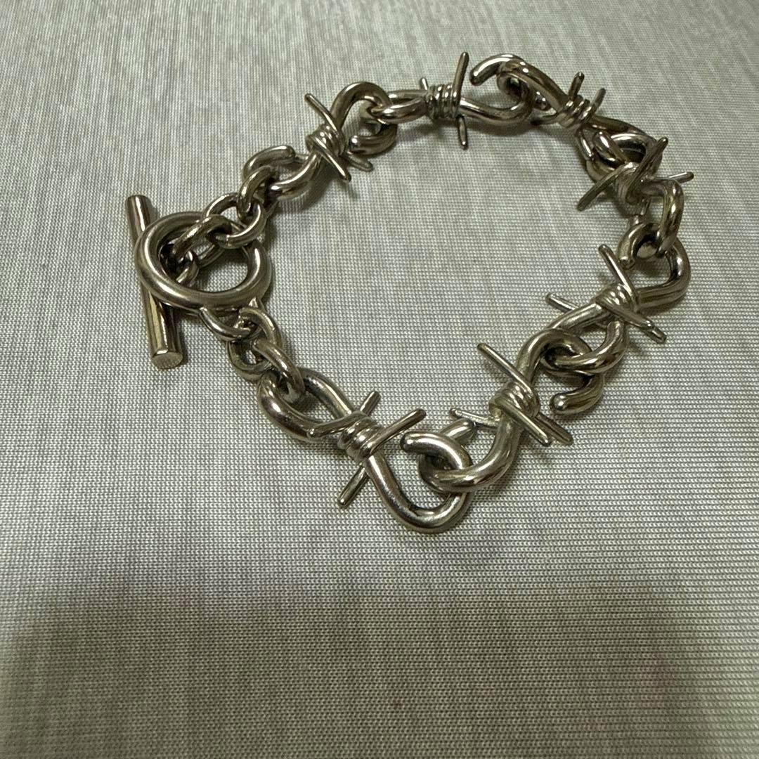 Silver UNDER COVER bracelet with barbed-wire-style chain links and a T-bar clasp laid on fabric.