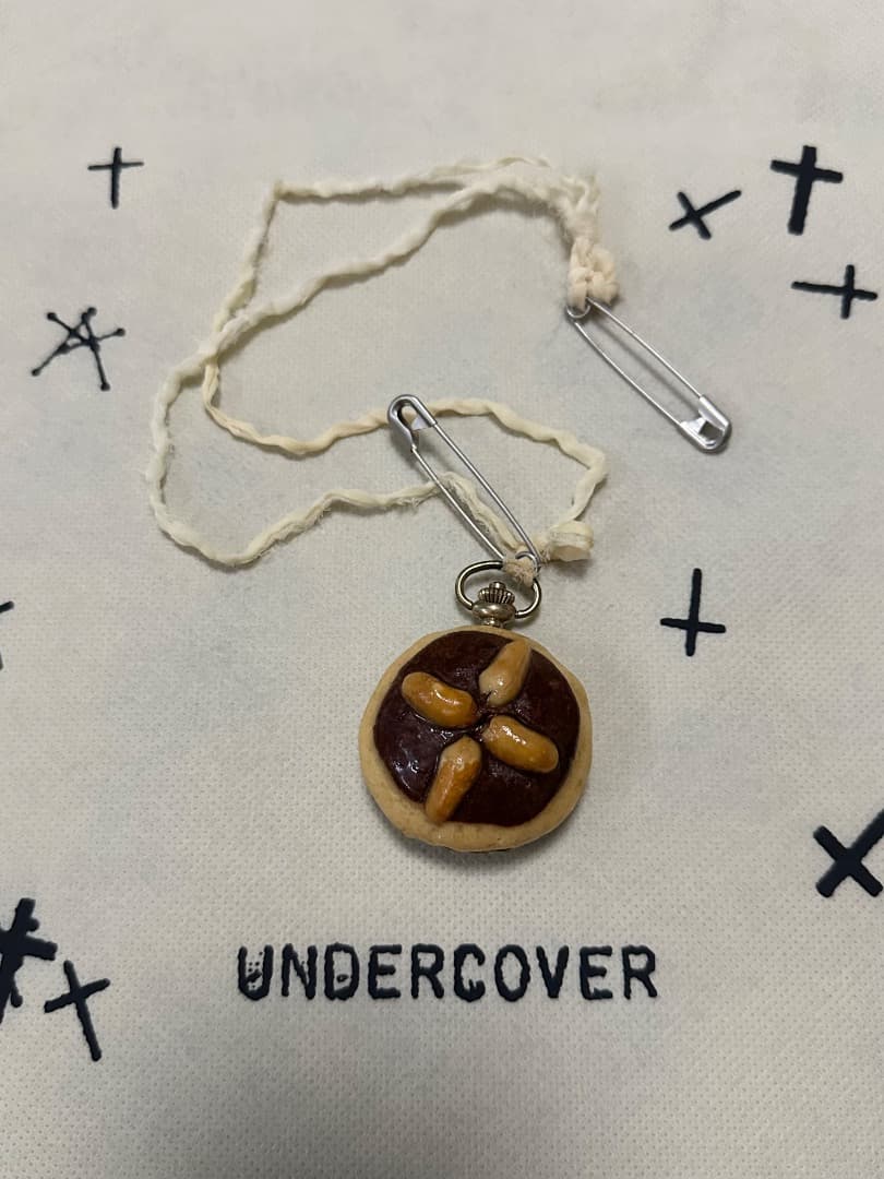 Round pocket watch with a brown center and four beige decorative elements, attached to a cord and safety pins, shown on a cloth printed with "UNDERCOVER".