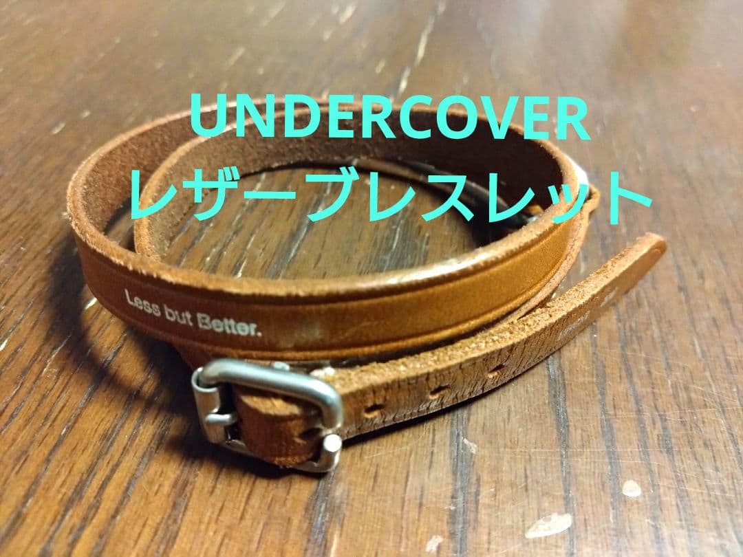 Brown cow leather bracelet coiled on a wooden surface, showing a small metal buckle and visible stamped text on the strap.