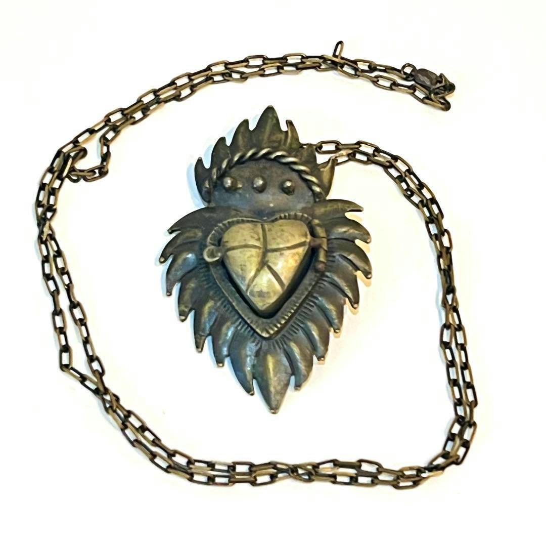 Photo of the アンダーカバー トラッシュ ロケットネックレス pendant showing a heart-shaped center with a flame-like surround on a metal chain.