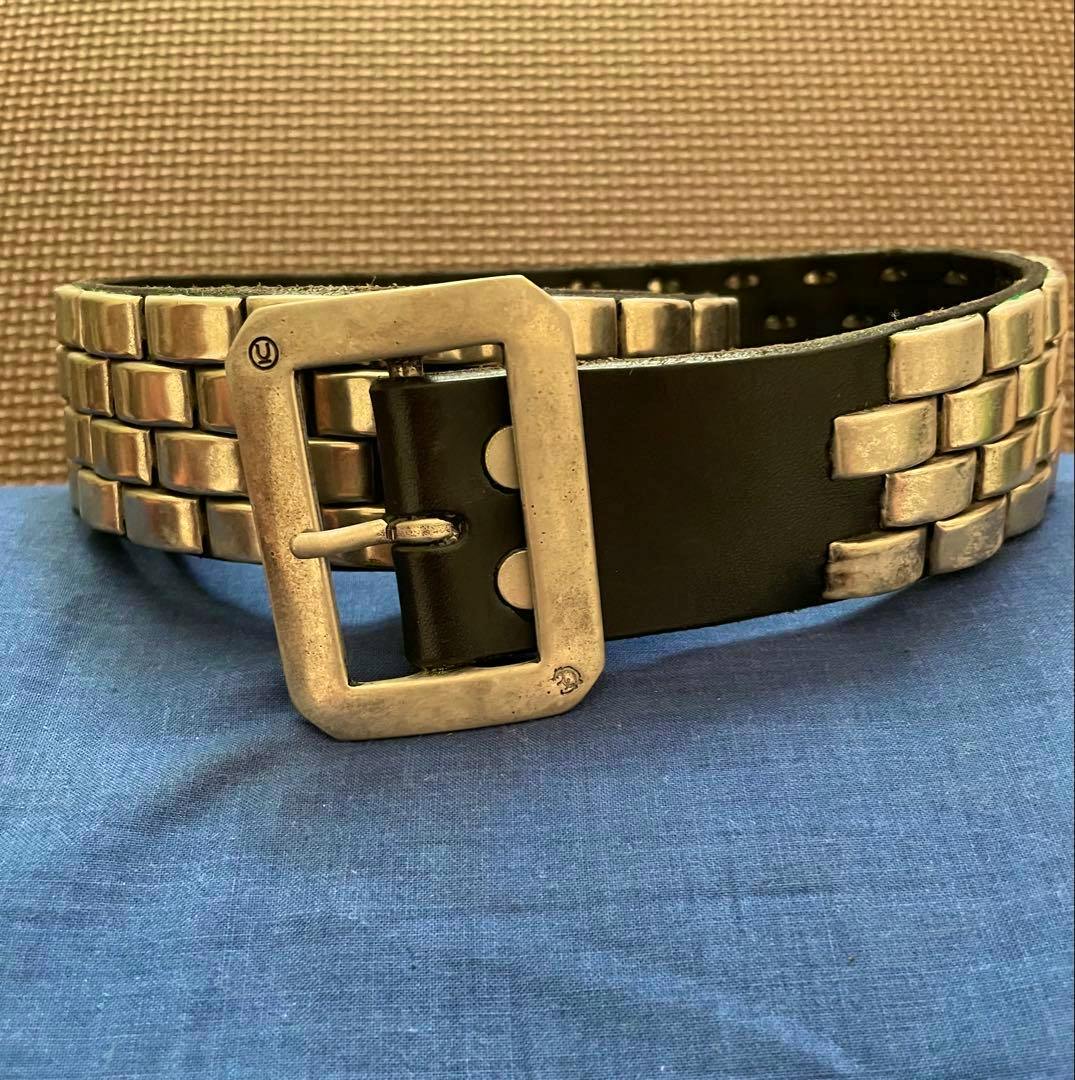 Black UNDERCOVER cow leather belt with three rows of metal studs and a rectangular aged-metal buckle displayed on a blue surface.