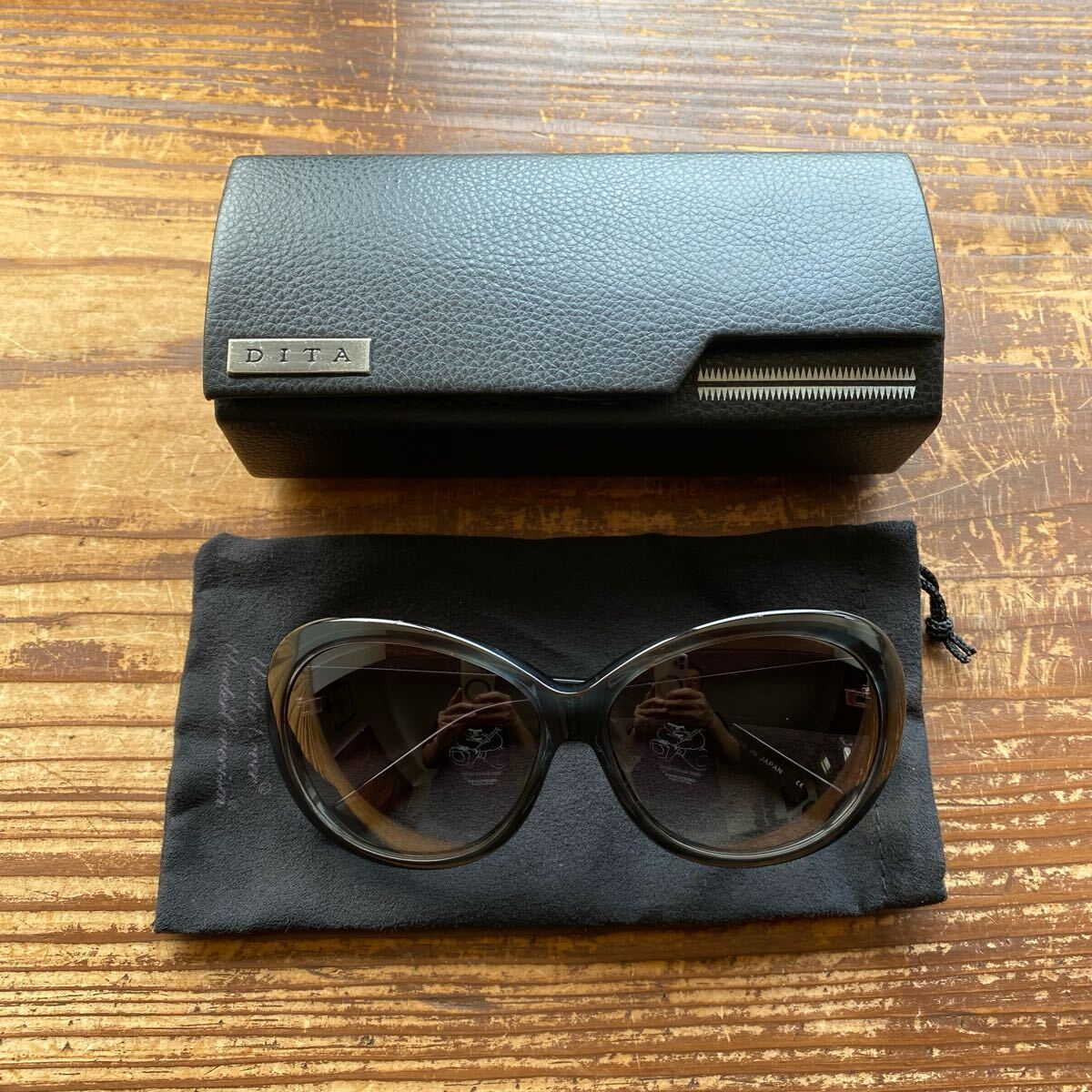 Black plastic sunglasses displayed on a soft pouch with a DITA hard case behind them.