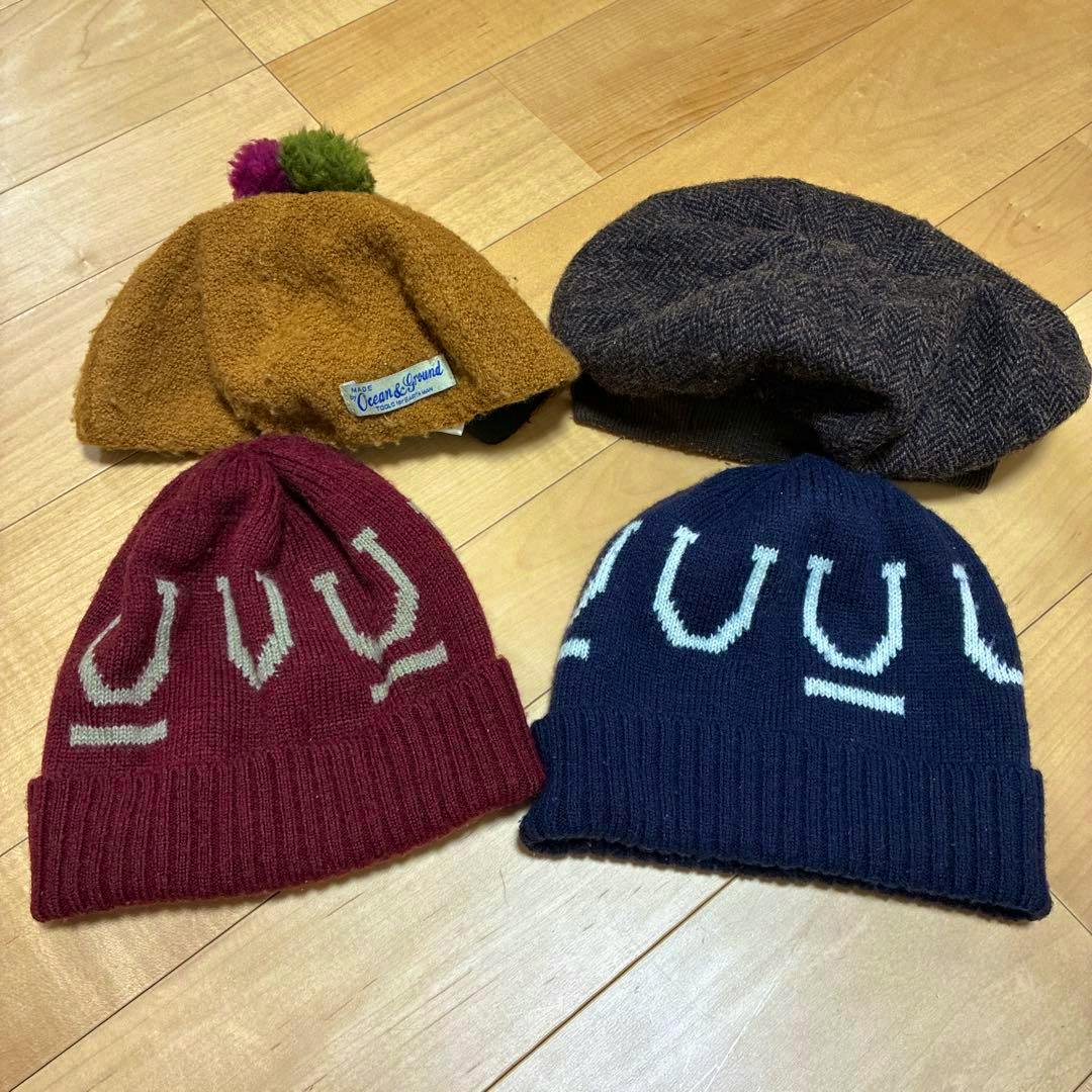 Four hats (berets and knit beanies) arranged on a wooden floor, including a brown fuzzy hat with a pompom.