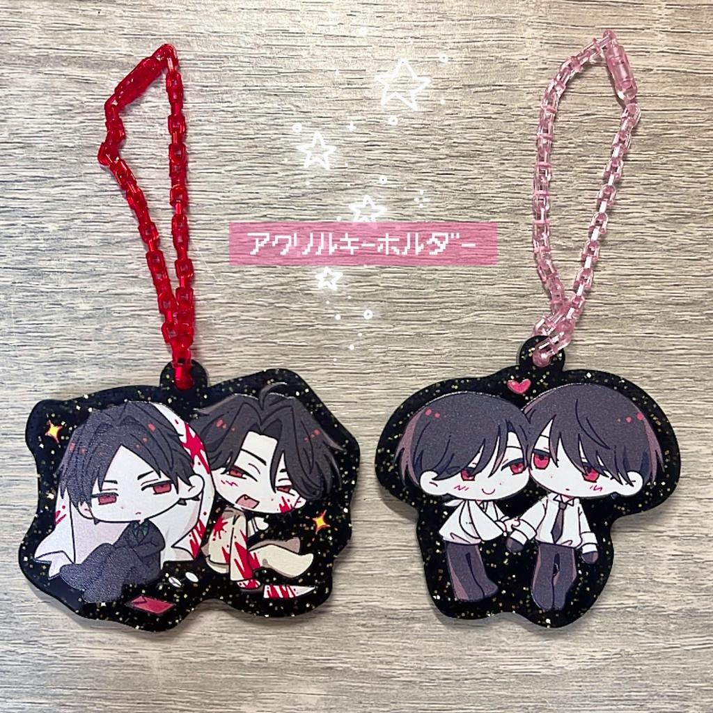 Two acrylic keychains with chibi character illustrations and colored chains placed on a wooden background.