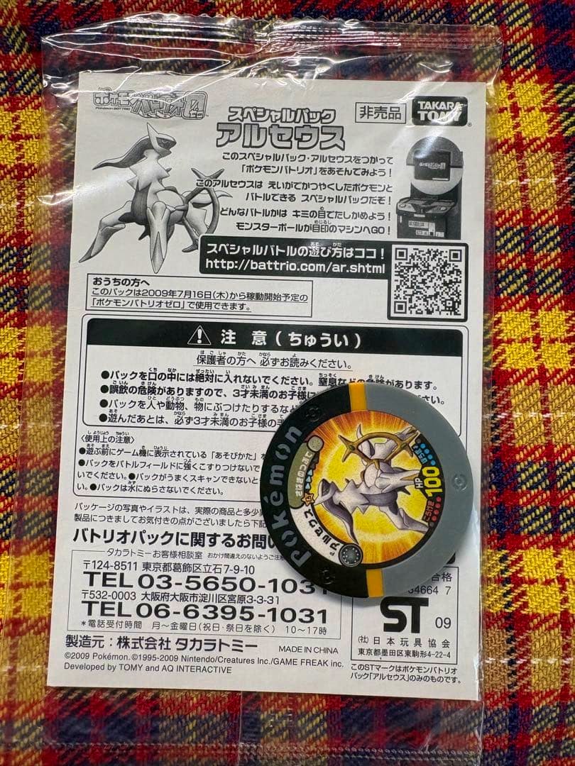 Sealed Pokémon card movie Arceus pack with Japanese insert and a round Arceus collectible disc on patterned fabric.