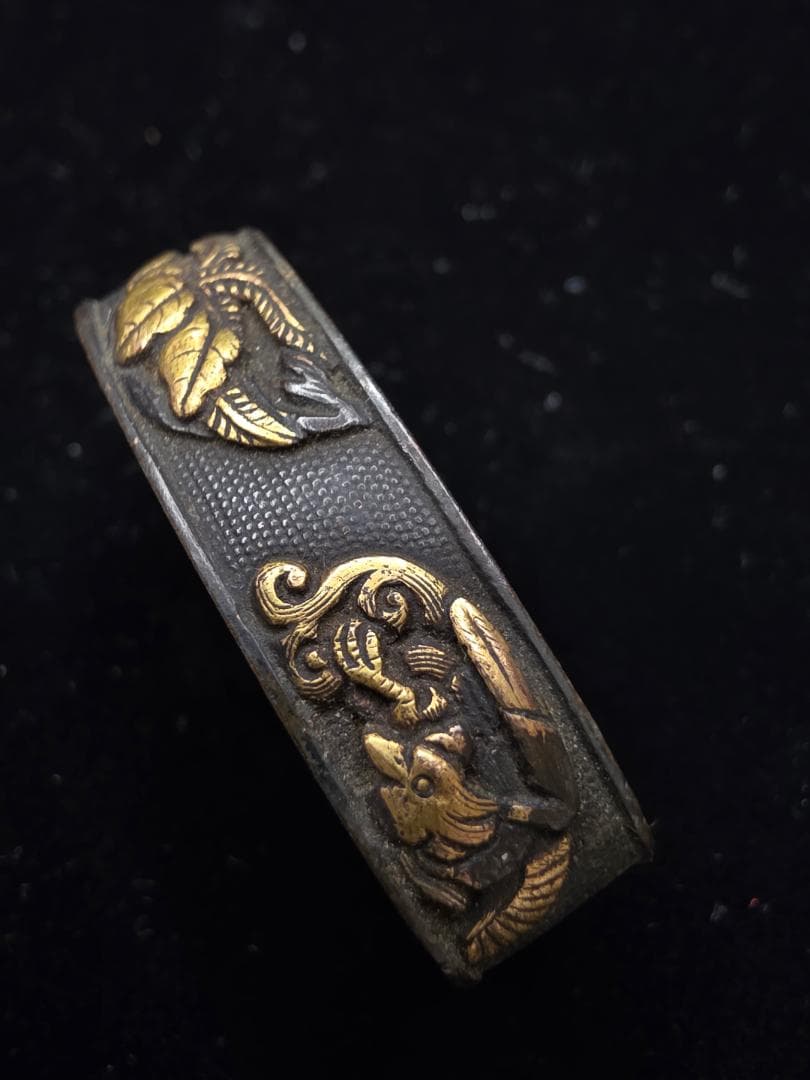 Close-up photo of a metal cufflink with gold floral and animal inlay on a textured dark background.