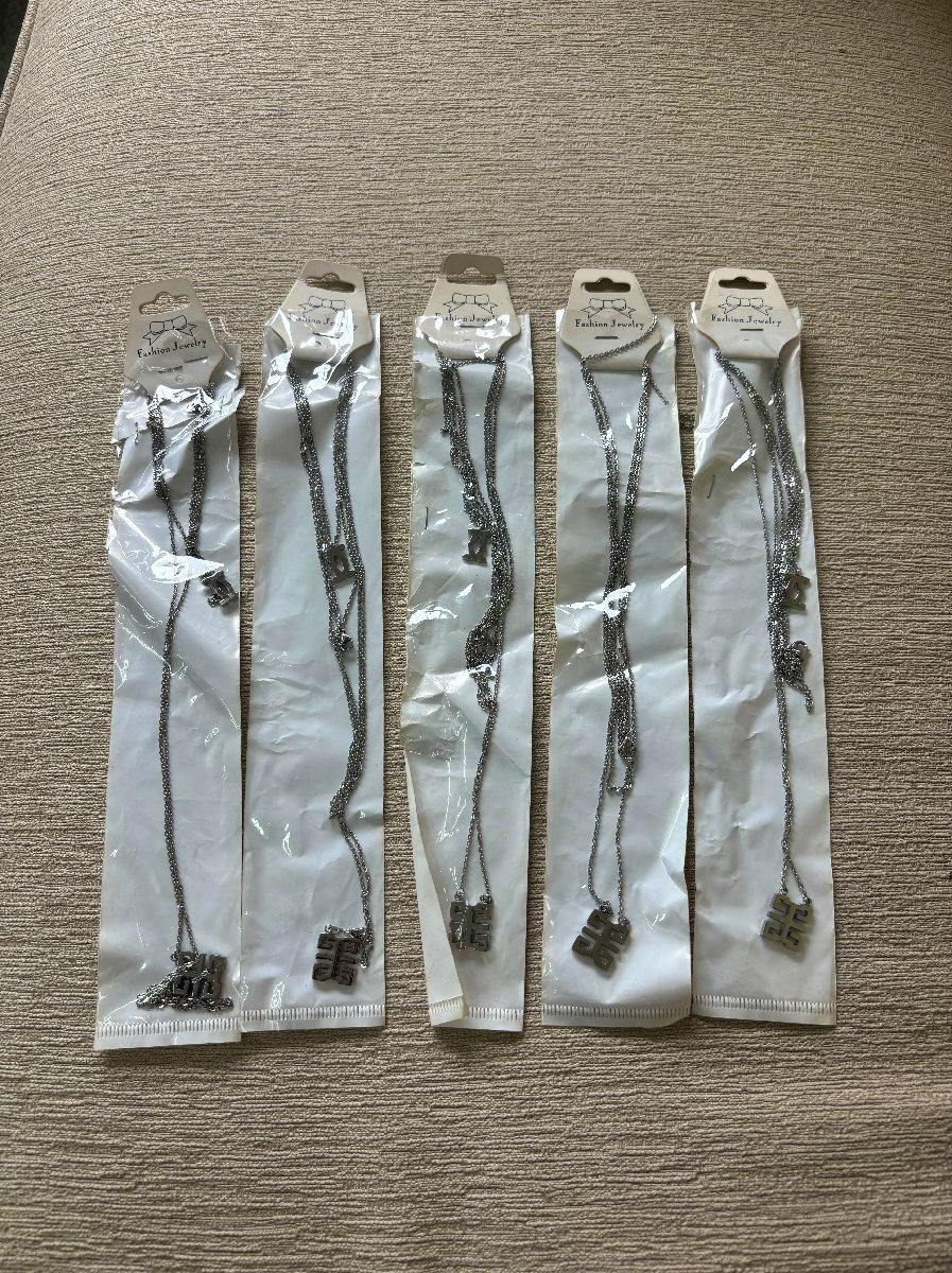 Five unused silver-tone necklaces in clear plastic packaging, each with a small pendant, laid side by side on a beige textured surface.