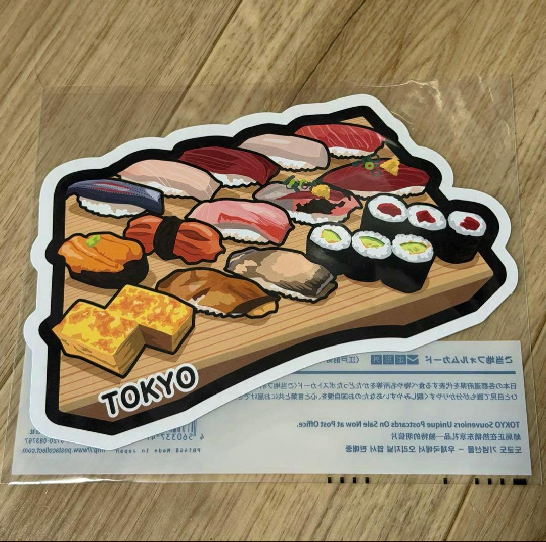 Shaped Tokyo souvenir postcard front showing an illustration of Edo-style sushi pieces on a wooden board, inside a plastic sleeve.