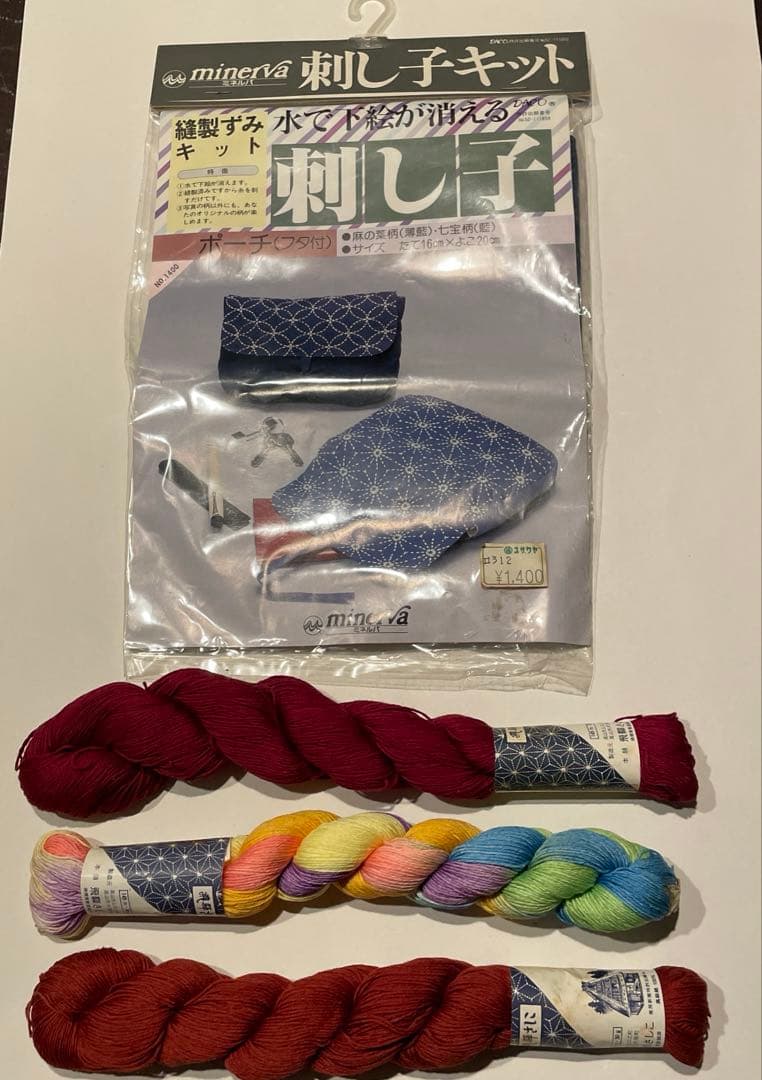 Packaged sashiko pouch kit with flap displayed above three skeins of sashiko thread on a white background.