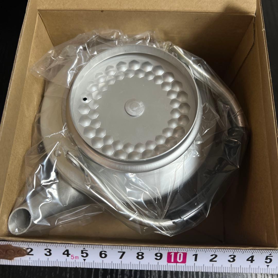 Top-down photo of a stainless steel honeycomb-pattern kettle wrapped in plastic inside a cardboard box with a measuring tape at the bottom.