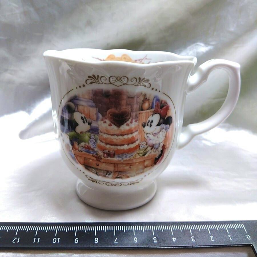 White ceramic Mickey & Minnie Collection cup showing a printed scene of Mickey and Minnie with a layered cake, with a ruler placed in front for scale.