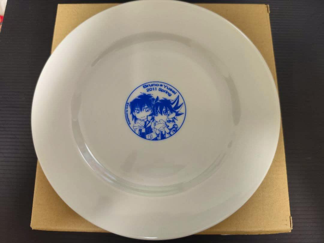 White ceramic plate with a small blue printed illustration of two chibi characters and text in the center, photographed on a cardboard box.