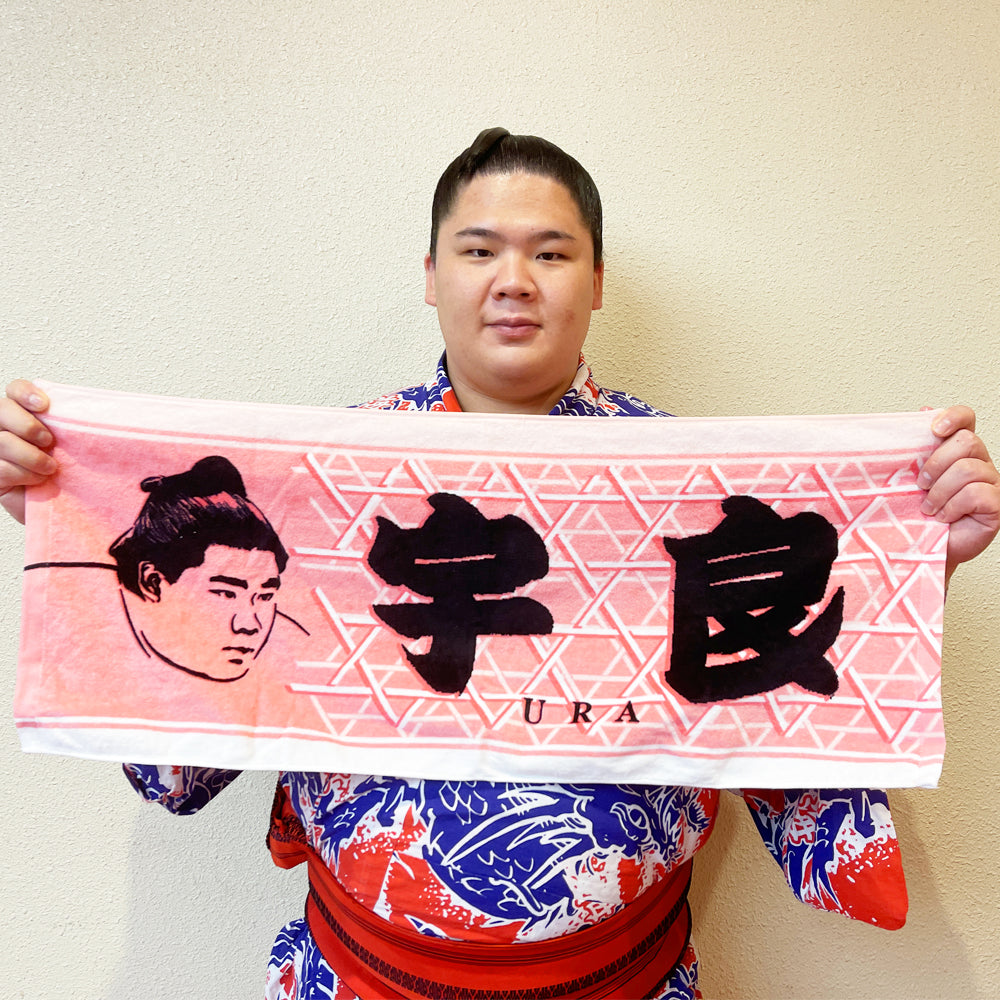 A person holds a pink towel printed with large black kanji characters reading "宇良" and a sumo wrestler portrait.