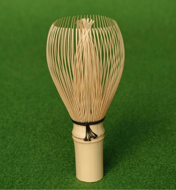 A bamboo tea whisk standing upright on a green surface, shown front-on.