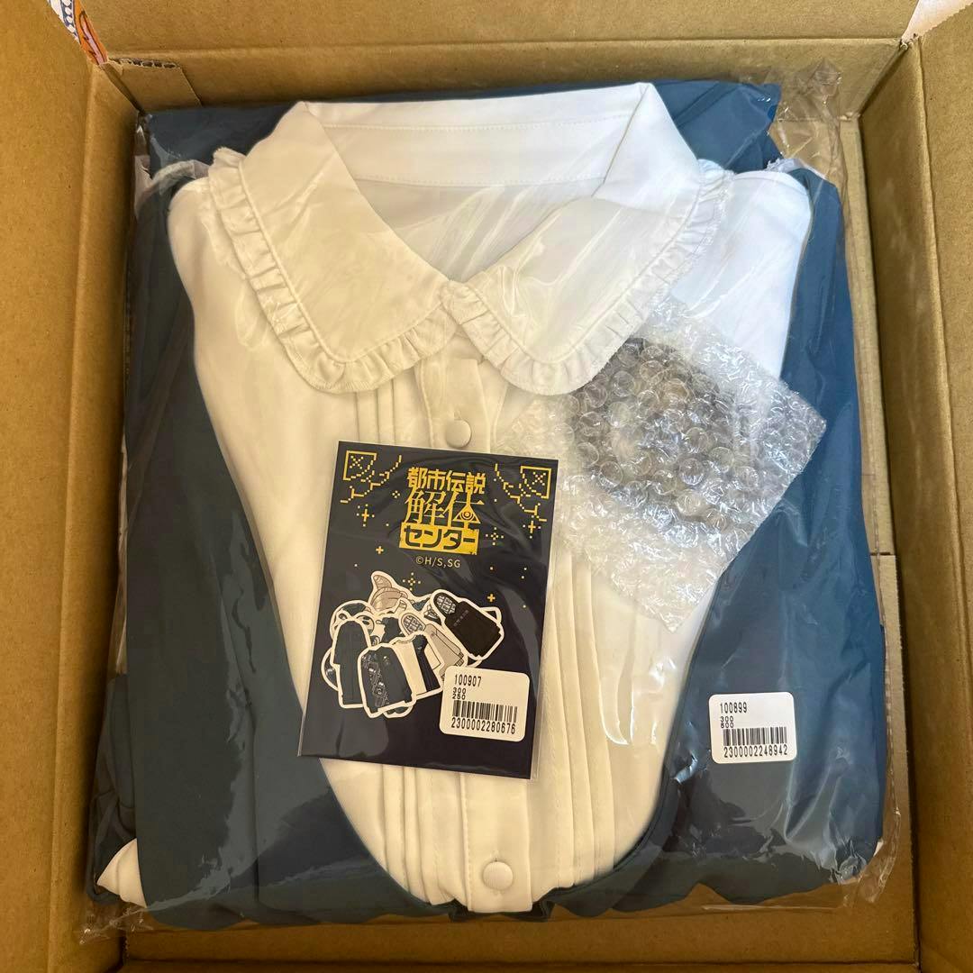 Unopened folded women's dress with a white ruffled collar and blue body sealed in plastic inside a cardboard box with a product tag visible.
