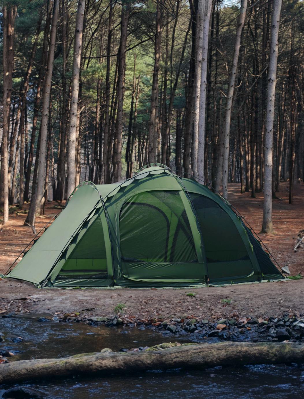 A green dome shelter tent set up on a forest floor beside a small stream.