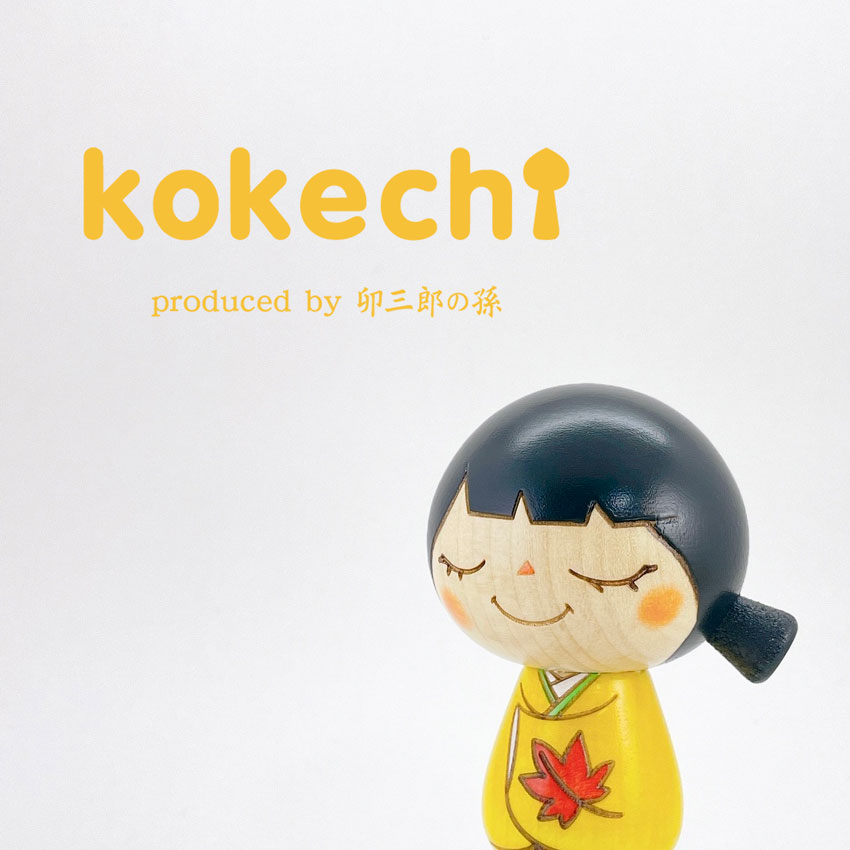 A wooden kokeshi doll wearing a yellow kimono decorated with a red maple leaf, smiling with closed eyes against a white background.