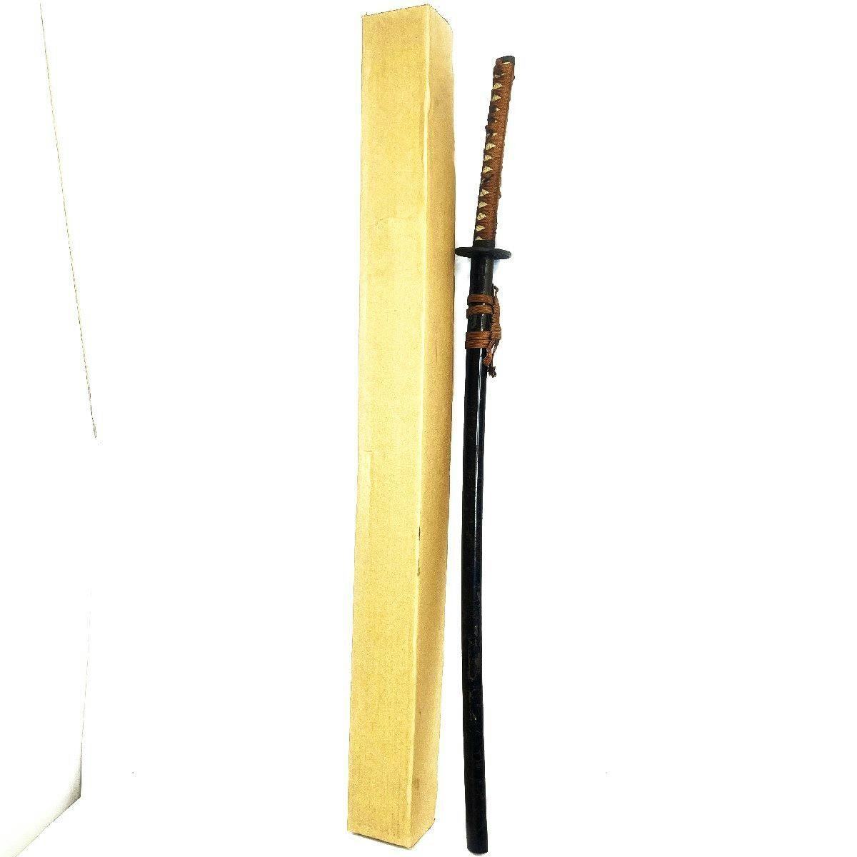 Imitation sword with a black scabbard and brown-wrapped handle standing next to a cardboard box on a white background.