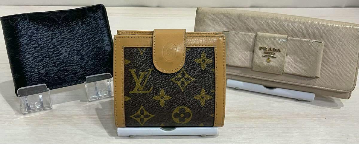 Three wallets displayed on stands: a black wallet at left, a Louis Vuitton monogram wallet center, and a beige Prada saffiano long wallet with a bow motif at right.