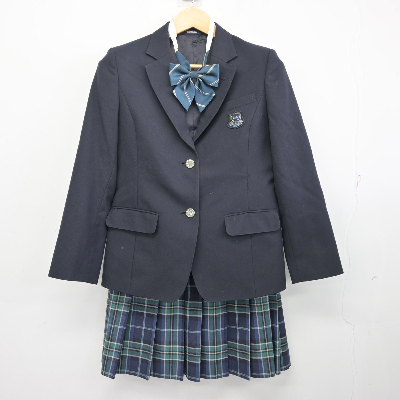 Navy blazer with school crest, striped bow tie, and pleated green-and-blue plaid skirt hanging on a hanger against a white background.