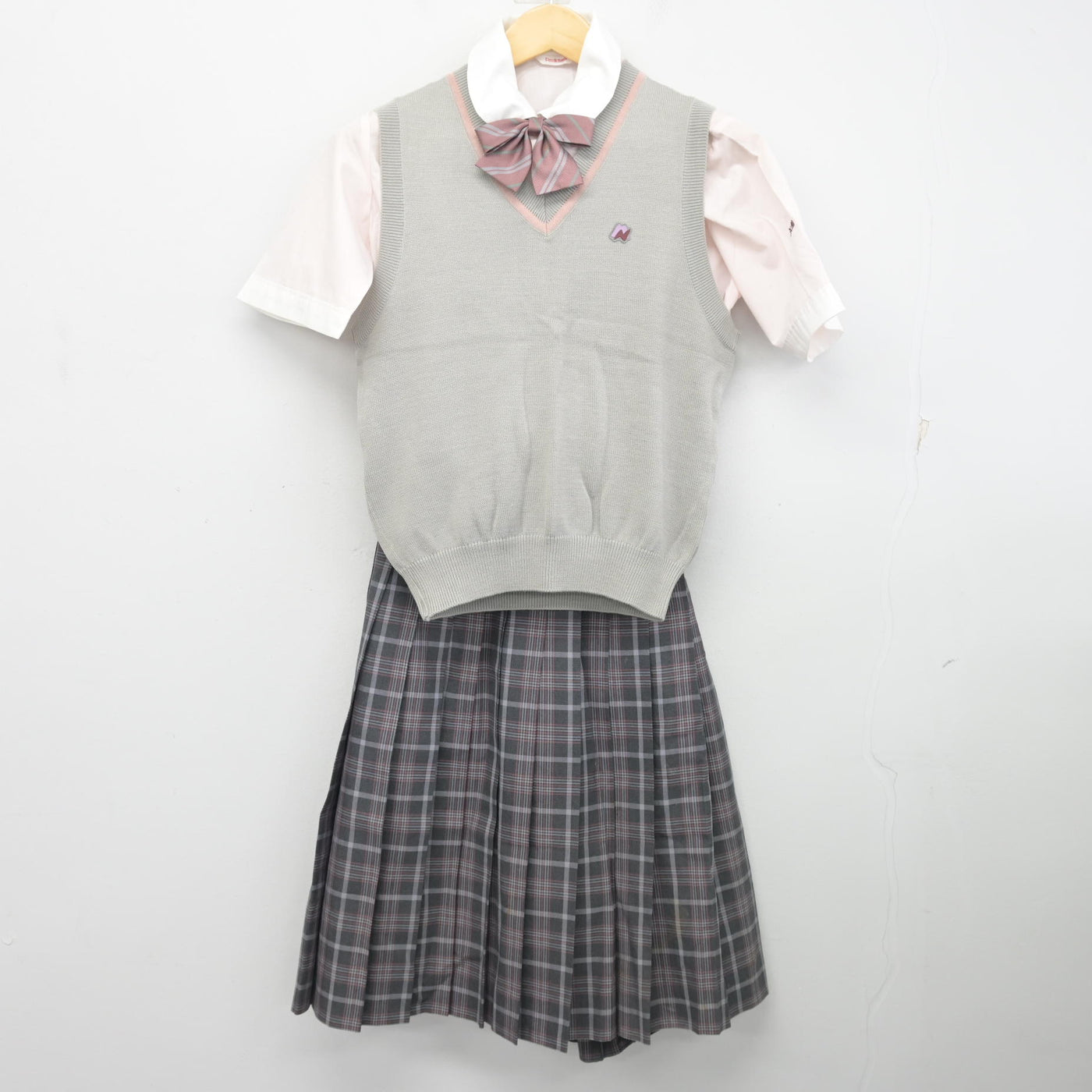 A school uniform set displayed on a hanger: a grey knit vest over a pale pink shirt with a bow and a grey plaid skirt against a white background.