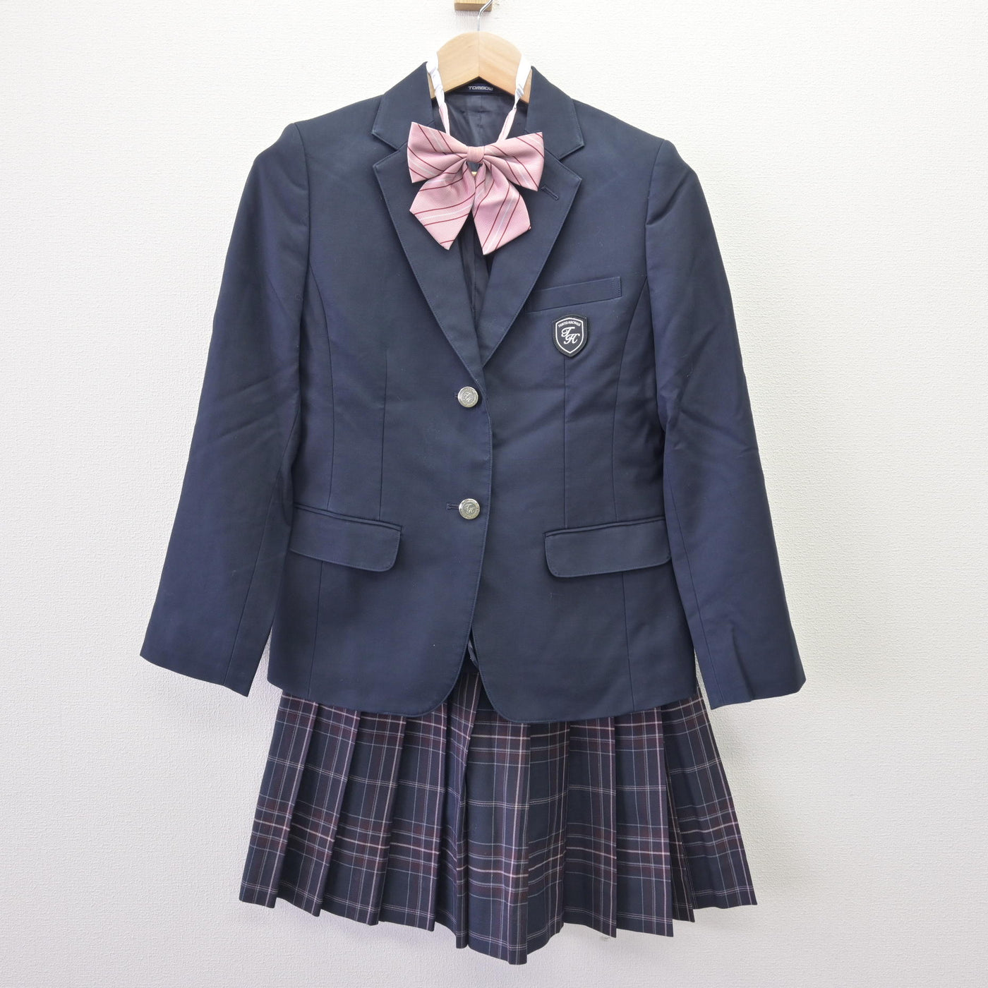 A navy blazer with a pink bow and a pleated plaid skirt hanging on a wooden hanger against a white background.