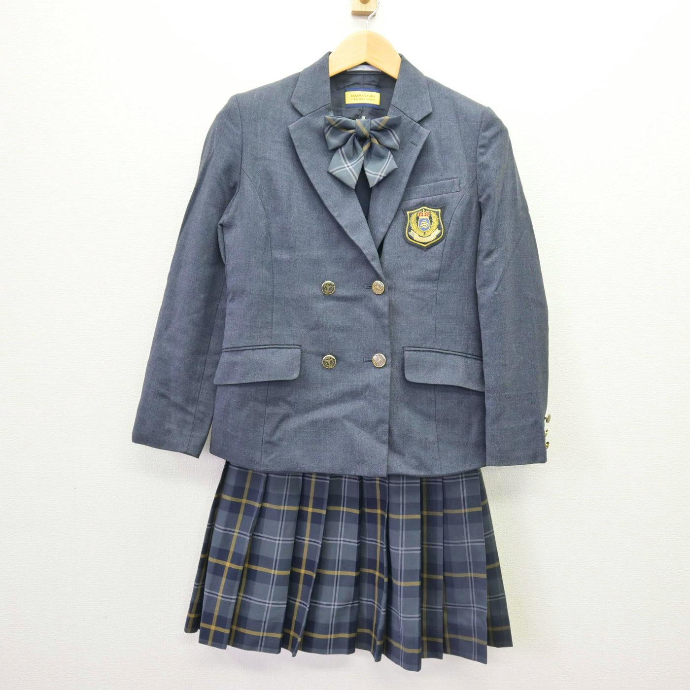 Gray school blazer with a crest, matching plaid pleated skirt, and a bow displayed on a hanger.