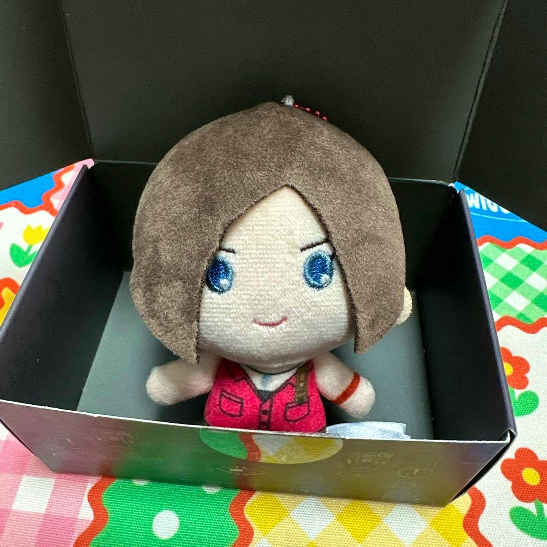 A USJ バイオハザード コレクタブルマスコット plush with brown hair, blue embroidered eyes, and a red outfit seated inside a black box.