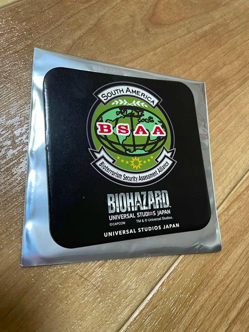 Sealed paper coaster showing a South America BSAA emblem and "BIOHAZARD UNIVERSAL STUDIOS JAPAN" text on a wooden floor.