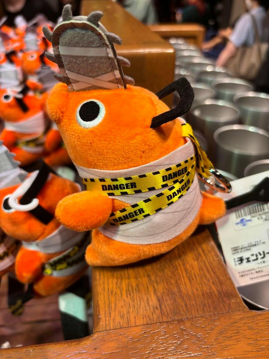 Orange Pochita plush keychain with a felt chainsaw on its head and yellow “DANGER” tape wrapped around it, shown on a wooden surface.