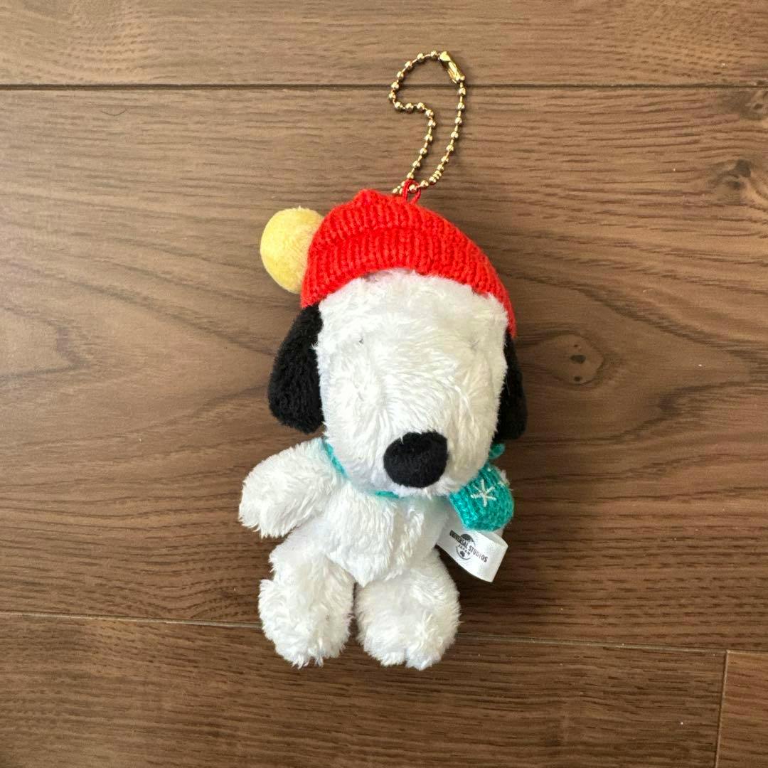 Front view of a Snoopy plush mascot wearing a red knit hat with a yellow pom-pom and a turquoise scarf, attached to a gold ball chain, on a wooden floor.
