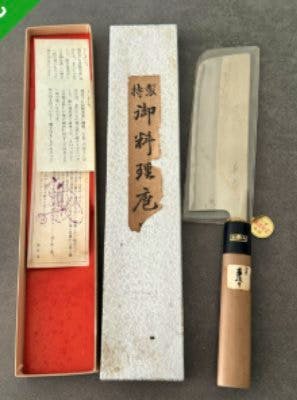 Thin-blade Japanese kitchen knife with wooden handle displayed next to its box and paperwork on a gray surface.