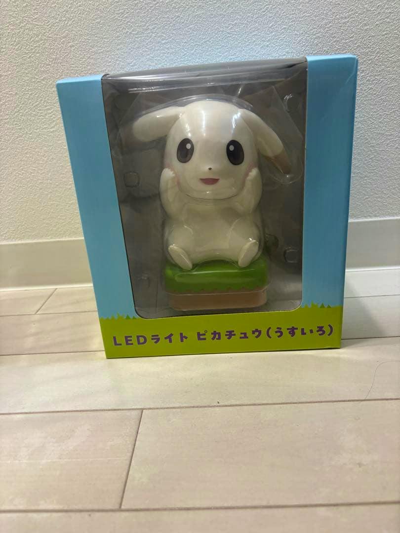 Packaged plastic LED light toy showing a pale Pikachu character sitting on a green base inside its box.