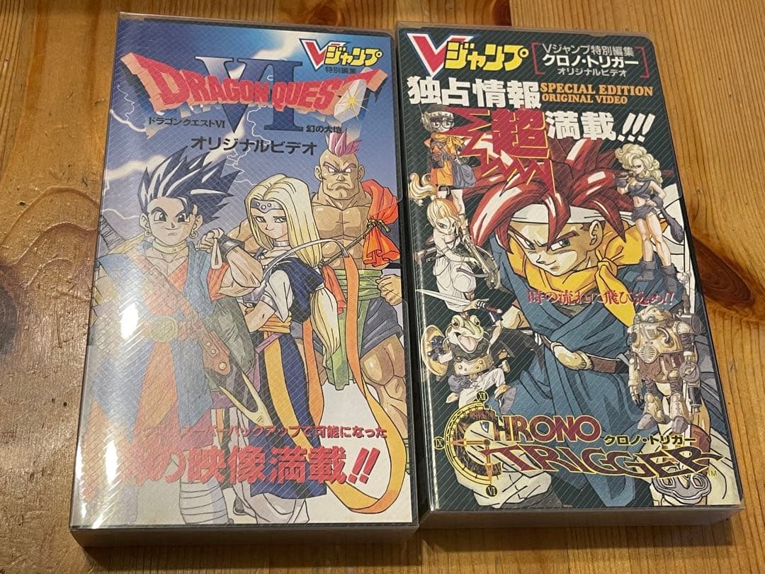 Two VHS tape cases side by side showing cover art for Vジャンプオリジナルビデオ (Chrono Trigger and Dragon Quest VI) on a wooden surface.